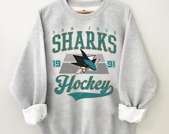 May include: A pale grey sweatshirt featuring the text "San Jose Sharks Hockey" in teal and white. The design includes a shark logo and the numbers "19" and "91". It has a crew neck and long sleeves with white cuffs.