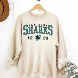 May include: A cream-colored sweatshirt with a vintage-style San Jose Sharks logo. The logo is in teal, black, and white and features the text "San Jose Sharks" and "Est. 1991".