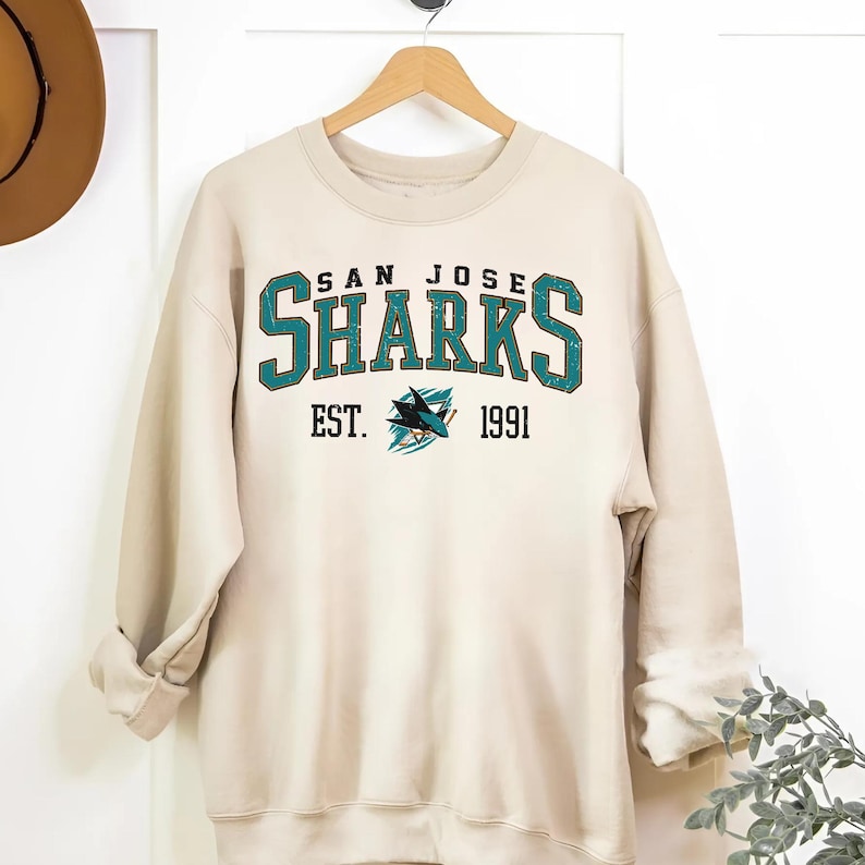 May include: A cream-colored sweatshirt with a vintage-style San Jose Sharks logo. The logo is in teal, black, and white and features the text "San Jose Sharks" and "Est. 1991".