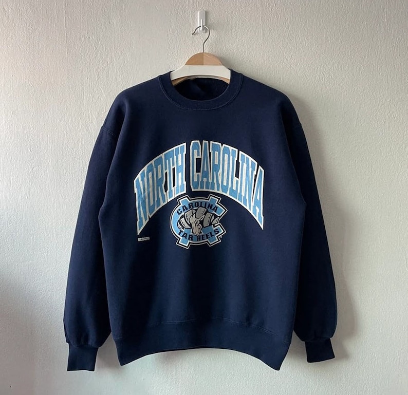 May include: A navy blue sweatshirt with a light blue graphic of the North Carolina Tar Heels logo. The logo features a fist with a basketball and the words "Carolina Tar Heels".