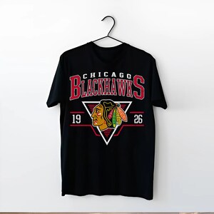 May include: Black t-shirt with a red, white, and black Chicago Blackhawks logo. The logo features a Native American headdress and the text "CHICAGO BLACKHAWKS" with the numbers "19" and "26" on either side.