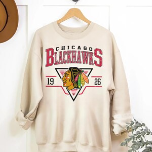 May include: A beige sweatshirt with a vintage-style graphic of the Chicago Blackhawks logo. The logo features a red and black Native American headdress with feathers and the text "Chicago Blackhawks" in red and black. The numbers "19" and "26" are on either side of the logo.