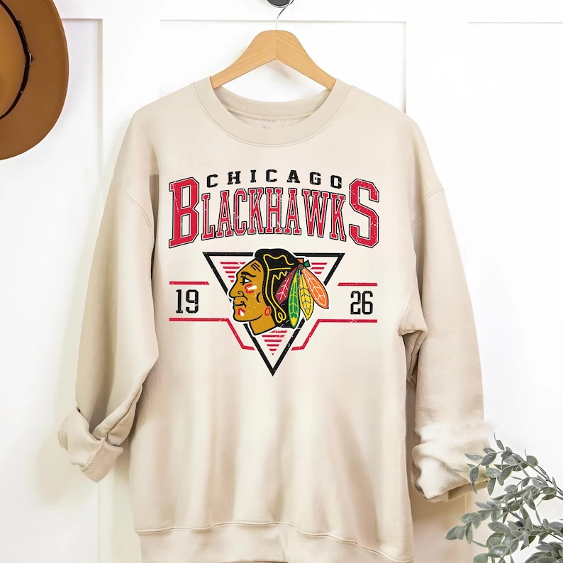 May include: A beige sweatshirt with a vintage-style graphic of the Chicago Blackhawks logo. The logo features a red and black Native American headdress with feathers and the text "Chicago Blackhawks" in red and black. The numbers "19" and "26" are on either side of the logo.