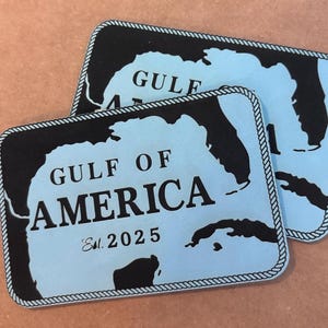 May include: Two rectangular, light blue plaques with black borders and text. The plaques read "GULF OF AMERICA" with a map outline and "Est. 2025". The borders have a rope-like design.