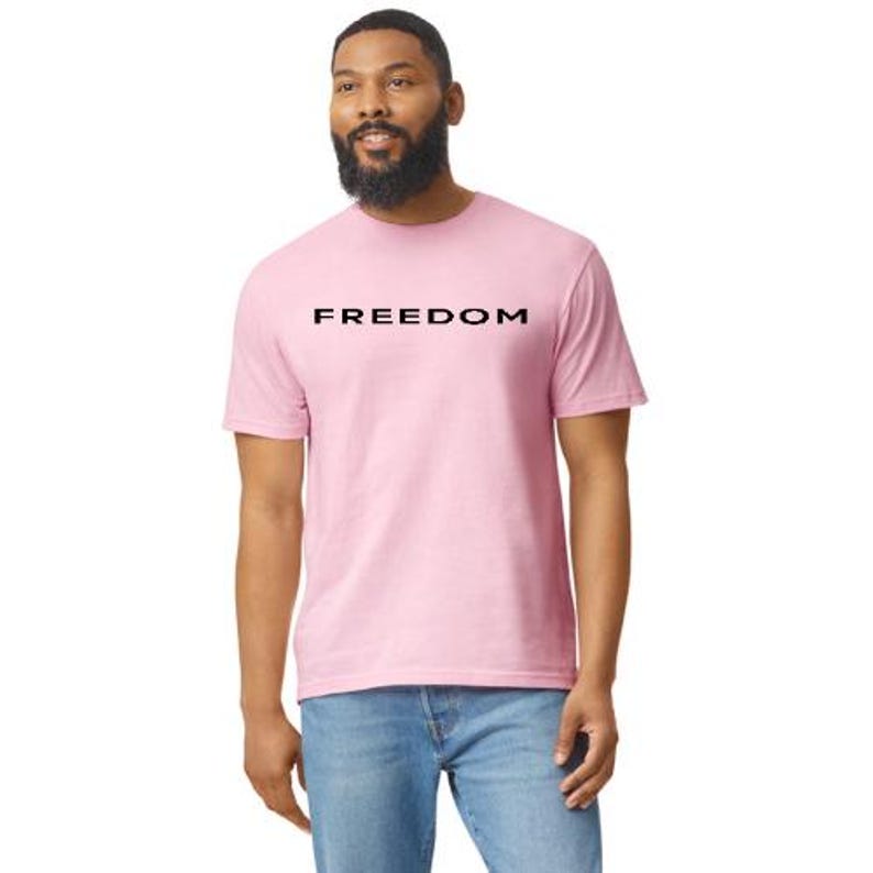 May include: A light pink t-shirt with the word "FREEDOM" printed in black across the chest. The shirt is short-sleeved and worn by a person wearing blue jeans.