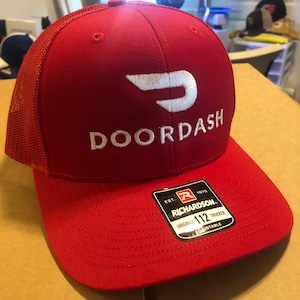 May include: Red and black trucker hat with a white DoorDash logo and a black Richardson 112 label. The hat is adjustable.
