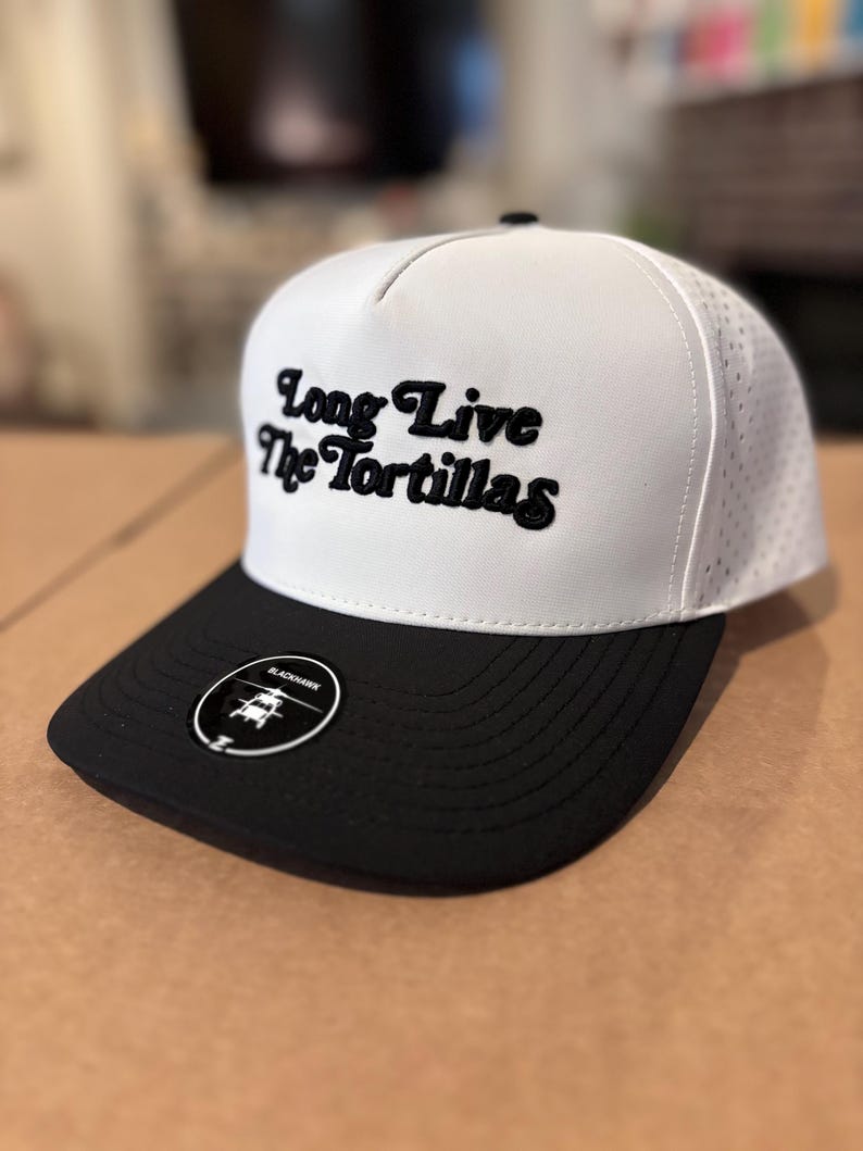May include: A white and black baseball cap with the text "Long Live The Tortillas" in black script. The cap has a black brim and a small circular tag with a logo.