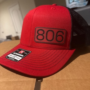 May include: Red and black trucker hat with a black leather patch that says "806" on it. The hat is adjustable and has a Richardson brand label.