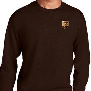 May include: A brown crew neck sweatshirt with the UPS logo on the upper left chest. The sweatshirt is worn by a person wearing blue jeans.