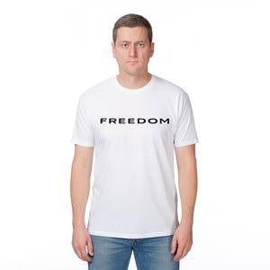 May include: A white t-shirt with the word "FREEDOM" printed in black, centered on the chest. The shirt is short-sleeved and has a crew neck. The person is wearing blue jeans.