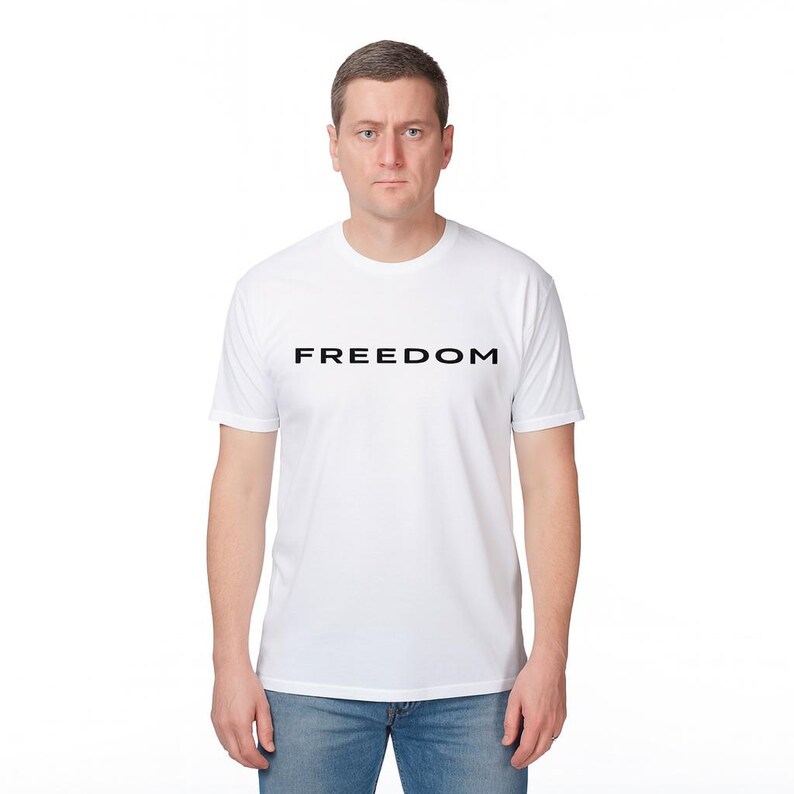 May include: A white t-shirt with the word "FREEDOM" printed in black, centered on the chest. The shirt is short-sleeved and has a crew neck. The person is wearing blue jeans.