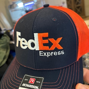 May include: A navy blue and orange trucker hat with the FedEx Express logo embroidered on the front. The hat has a red and white label that says "EST. 1970 RICHARDSON® ORIGINAL 112 TRUCKER ADJUSTABLE".