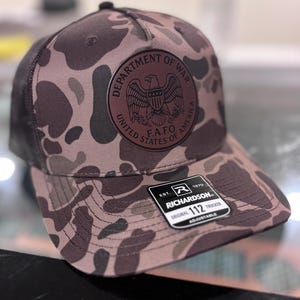 May include: A camouflage trucker hat with a brown and black pattern. The hat features a circular patch with an eagle emblem and the text "DEPARTMENT OF WAR UNITED STATES OF AMERICA F.A.F.O." The hat has a black mesh back and a curved brim.
