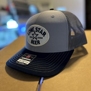 May include: A gray and black trucker hat with a white patch that says "Lone Star Beer 1984 2024" with a blue star in the center. The hat has a black mesh back and a Richardson 112 Trucker label.
