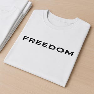 May include: White t-shirts with the word "FREEDOM" printed in black. The shirts are folded and stacked on a light brown surface. The t-shirts have a crew neck design.