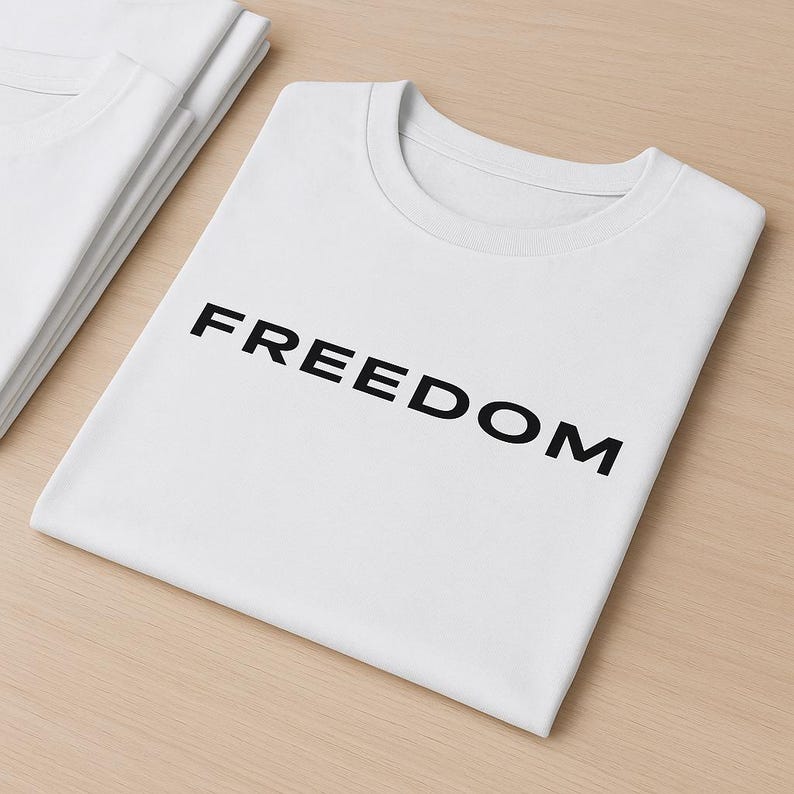 May include: White t-shirts with the word "FREEDOM" printed in black. The shirts are folded and stacked on a light brown surface. The t-shirts have a crew neck design.