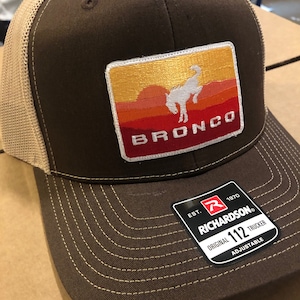 May include: Brown and tan trucker hat with a brown patch that says "BRONCO" in white letters. The patch has a white silhouette of a bucking bronco horse against a sunset background. The hat has a black adjustable strap with a white label that says "EST. 1970 RICHARDSON ORIGINAL 112 TRUCKER ADJUSTABLE".