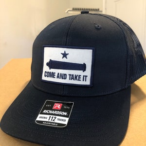 May include: A navy blue trucker hat with a white patch featuring a cannon and a star with the text "Come and Take It". The hat has a black mesh back and a red and black label that reads "Richardson Original 112 Trucker Adjustable".