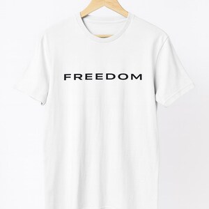 May include: White t-shirt with the word "FREEDOM" printed in black, displayed on a wooden hanger. The shirt has a crew neck and short sleeves, and is made of a soft, likely cotton, material.