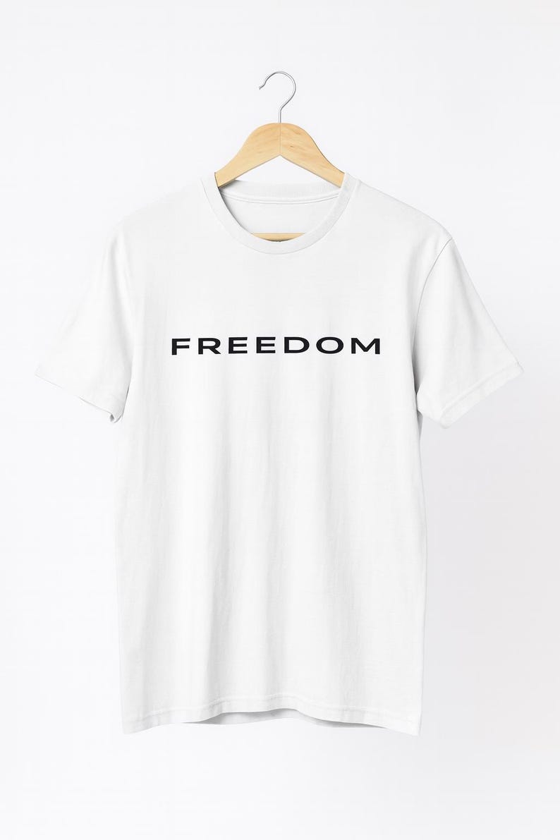 May include: White t-shirt with the word "FREEDOM" printed in black, displayed on a wooden hanger. The shirt has a crew neck and short sleeves, and is made of a soft, likely cotton, material.