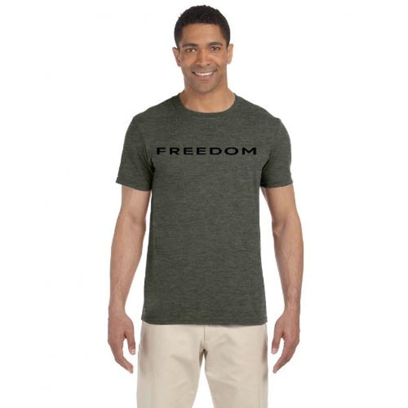 May include: A short-sleeved, olive green t-shirt with the word "FREEDOM" printed in black across the chest. The shirt is worn by a person, and paired with khaki pants. The person is smiling.