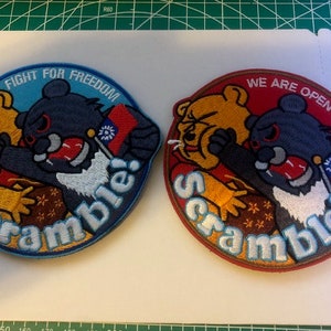 May include: Two embroidered patches with a cartoon bear and a black and gray creature. The left patch is blue and says "Fight for Freedom Scramble!" The right patch is red and says "We are Open 24/7 Scramble!"
