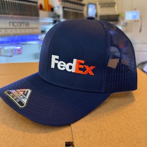 May include: A navy blue trucker cap featuring the white and orange FedEx logo embroidered on the front. The cap has a solid front panel, a curved brim, and a mesh back. A small sticker with the Pacific Headwear logo is attached to the brim.