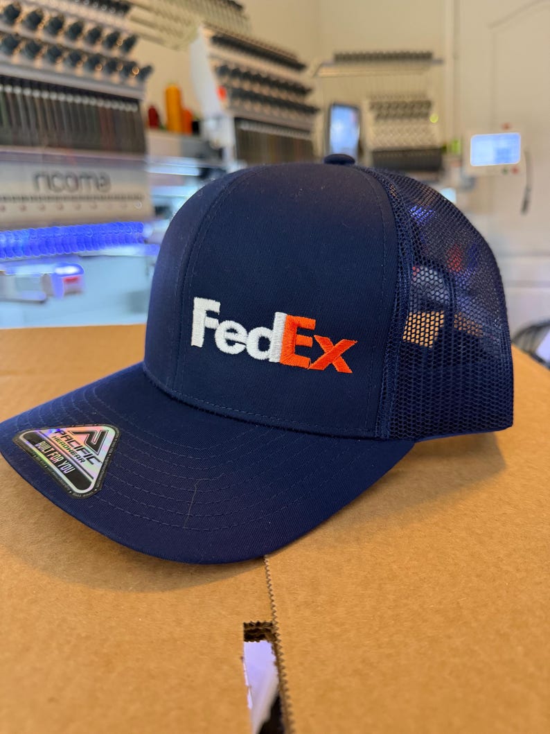 May include: A navy blue trucker cap featuring the white and orange FedEx logo embroidered on the front. The cap has a solid front panel, a curved brim, and a mesh back. A small sticker with the Pacific Headwear logo is attached to the brim.