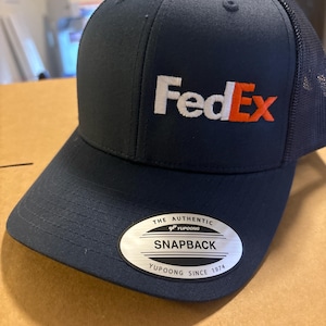 May include: A dark navy blue trucker cap with a mesh back and a structured front panel featuring the FedEx logo embroidered in white and orange. A silver oval sticker on the brim reads The Authentic Yupoong Snapback Yupoong Since 1974.
