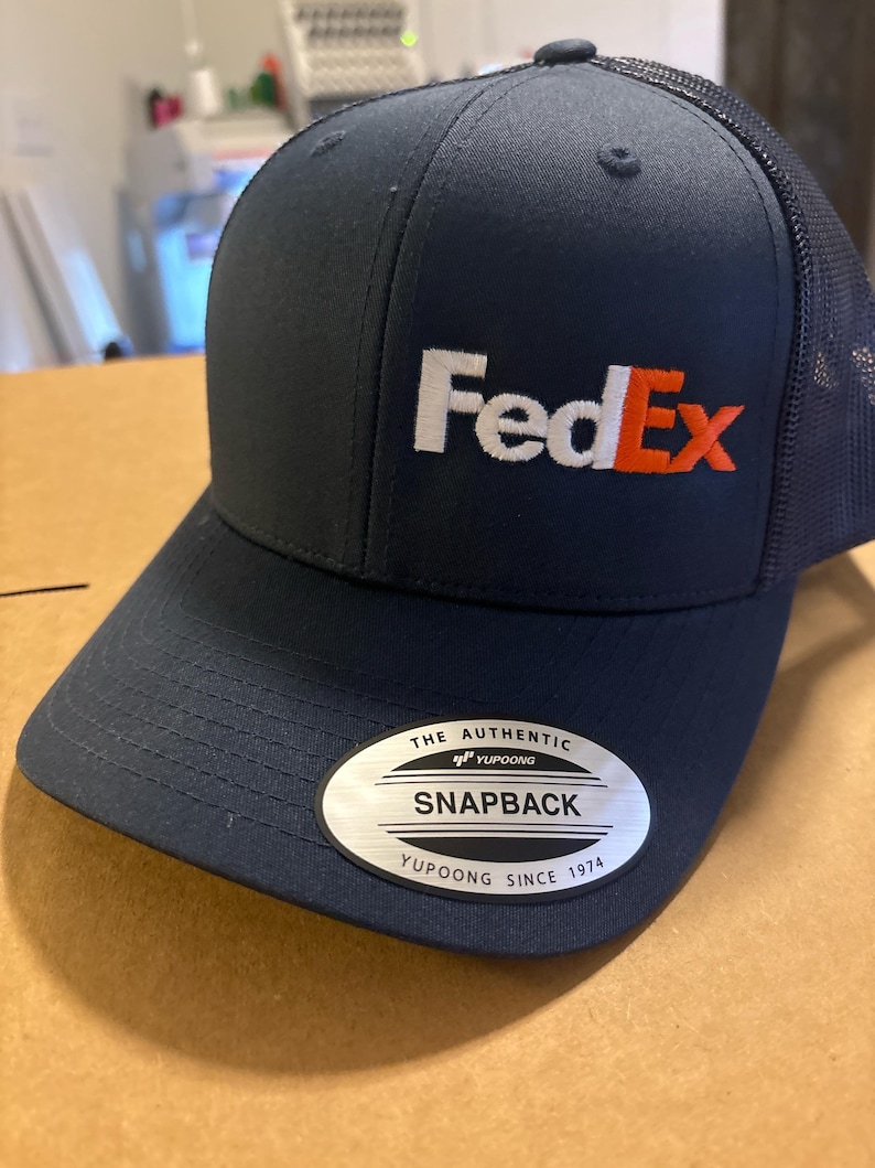 May include: A dark navy blue trucker cap with a mesh back and a structured front panel featuring the FedEx logo embroidered in white and orange. A silver oval sticker on the brim reads The Authentic Yupoong Snapback Yupoong Since 1974.