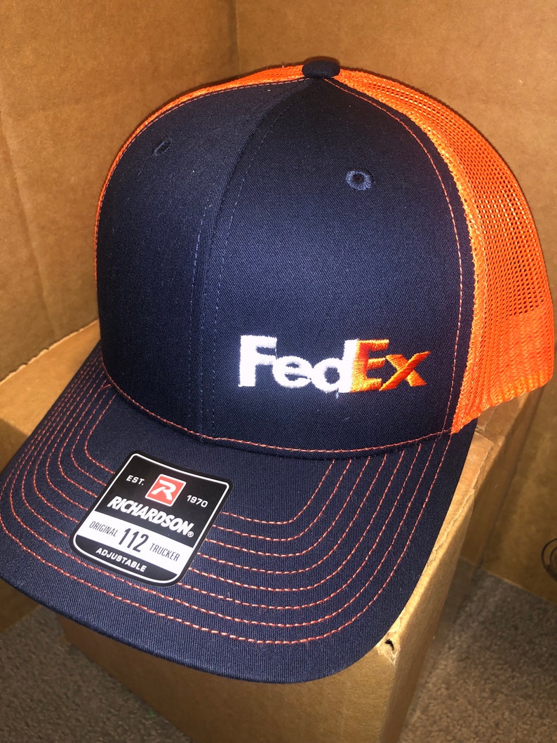 May include: A navy blue trucker hat with a bright orange mesh back and orange stitching on the brim. The front features the white and orange FedEx logo. A black Richardson 112 adjustable sticker is attached to the brim, displaying the brand name.