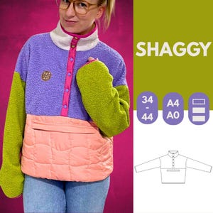 Puede incluir: Un jersey de forro polar con bloques de color y un bolsillo acolchado color melocotón. La parte superior presenta una sección superior morada, mangas verdes y un cuello blanco y rosa. La palabra "SHAGGY" se muestra en texto verde. Se enumeran las opciones de tamaño.