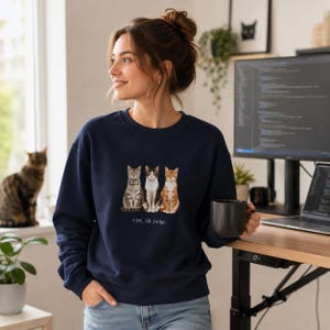 May include: A navy blue crewneck sweatshirt featuring a graphic of three cats sitting side-by-side with the text "Ctrl. Alt. Judge." printed underneath. The cats are depicted in brown, grey, and white tones, and the sweatshirt has long sleeves.