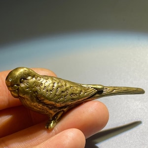 May include: A small, gold-coloured metal figurine of a parakeet. The figurine is detailed with feathers and has a long tail.