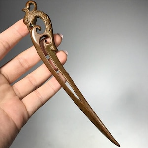 May include: A bronze hair stick with an intricate design featuring a bird at the top. The stick is long and slender, with a pointed tip.