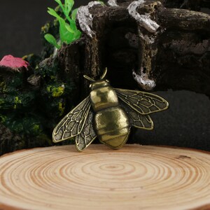May include: A gold-coloured metal bee charm with detailed wings. The bee is sitting on a wooden surface.