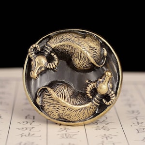 May include: A round, brass ornament with two stylized oxen facing each other. The oxen are in a yin-yang style design, with one facing up and the other facing down. The ornament has a dark brown patina.