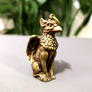 May include: A small, gold-coloured metal figurine of a griffin, a mythical creature with the head and wings of an eagle and the body of a lion. The figurine is sitting on a white surface.