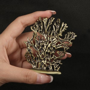 Op de afbeelding: Een goudkleurig metalen sculptuur van een gestileerde koraaltak. Het sculptuur is gedetailleerd met ingewikkelde patronen en texturen.