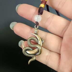 Puede incluir: Un collar con un colgante de serpiente dorado, con un diseño de serpiente enroscada detallado. El collar está en un cordón marrón con una cuenta blanca.