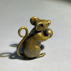 Puede incluir: Una pequeña figurita de metal dorado de un ratón sosteniendo un objeto redondo en sus patas. El ratón tiene una cola enroscada y detalles elaborados.