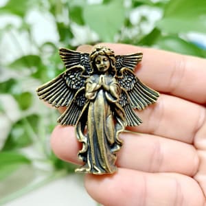 Może przedstawiać: Złota metalowa figurka anioła z rozłożonymi skrzydłami i złożonymi rękami w modlitwie. Anioł ma na sobie długą, falującą szatę i koronę na głowie.