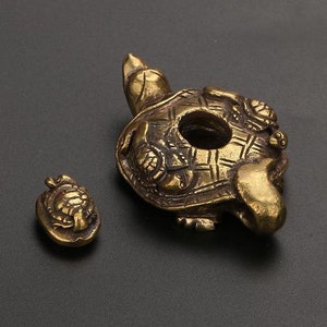 Puede incluir: Dos amuletos de tortuga de color dorado. El amuleto más grande es una tortuga detallada con una abertura redonda en el centro. El amuleto más pequeño es un diseño de tortuga simple.