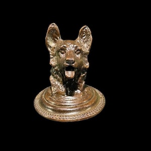 May include: A bronze sculpture of a dog's head with an open mouth, mounted on a circular base with a beaded edge.
