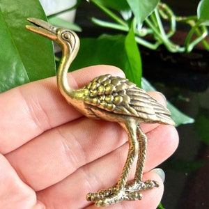 Pode incluir: Uma pequena figura de metal dourado de uma grulha com detalhes precisos, incluindo um pescoço longo, pernas longas e um bico. A figura está em pé sobre uma mão.