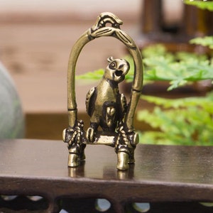 May include: A brass figurine of a parrot perched on a swing. The swing is made of a stylised bamboo design. The parrot is detailed with a textured body and a bright yellow beak.