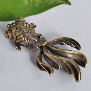 May include: A gold-coloured metal figurine of a fish with a detailed, scaly body and a long, flowing tail. The fish is facing to the right, and its tail is spread out behind it.