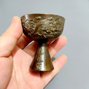 May include: A small, bronze cup with a dragon design and Chinese characters on the base. The cup is on a stand and has a rounded shape.
