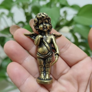 May include: A small, gold-colored metal figurine of a cherub with wings, holding an arrow. The cherub is standing on a round base.