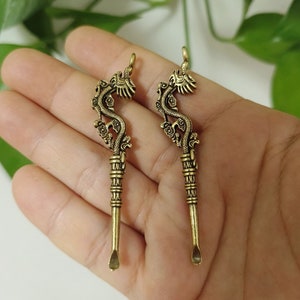 Puede incluir: Dos colgantes de metal color bronce. Cada colgante presenta un diseño de dragón en la parte superior, con un cuerpo curvado y un pequeño extremo en forma de cuchara. Los colgantes se muestran contra una mano.